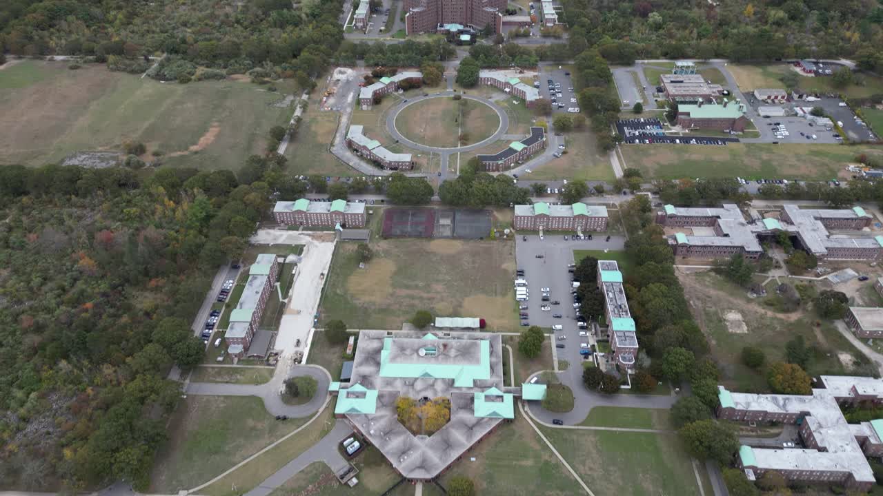 Aerial drone view of a large medical campus in Brentwood, Suffolk County, Long Island, NY, showcasing multi-wing architecture, structured parking, open grounds, and surrounding landscape in clear day