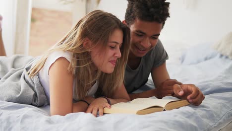 happy smiling multi ethnic couple lying in bed reading a book together at home in bedroom. slow motion shot