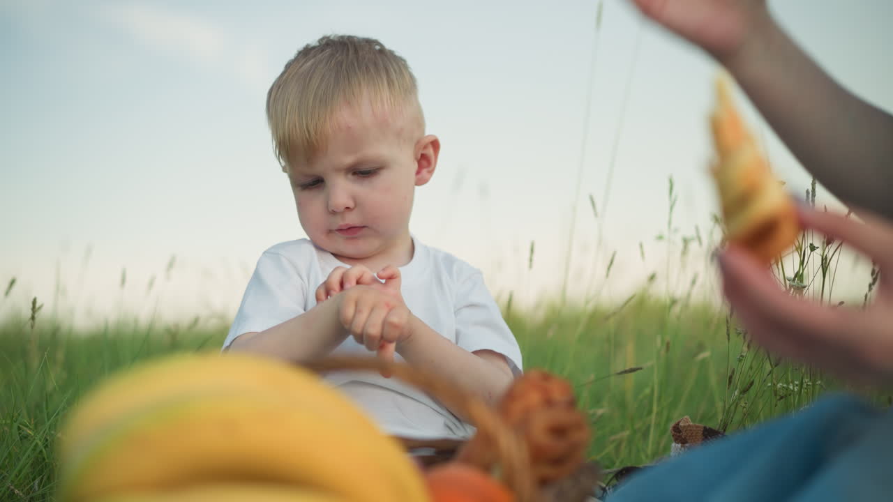 A little boy wearing a white shirt sits in a grassy field holding a snack. After taking a bite, he hands the snack to his mother, who is partially visible, sharing a sweet, nurturing moment outdoors