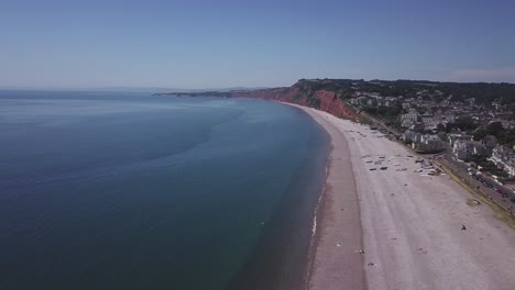 Weite-Luftaufnahme-Eines-Blauen-Meeres,-Eines-Kiesstrandes,-Rostroter-Trias-Klippen-Und-Der-Ruhigen-Stadt-Budleigh-Salterton