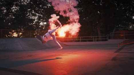 young guy is holding glowing red signal flare, jumping and spinning while performing breakdance on pump track of skatepark. slow motion