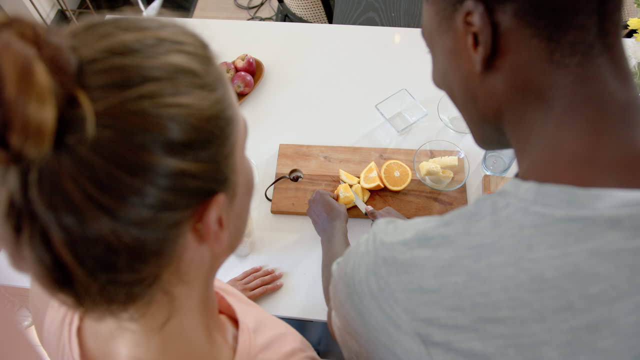 Diverse couple slicing oranges at kitchen counter, at home