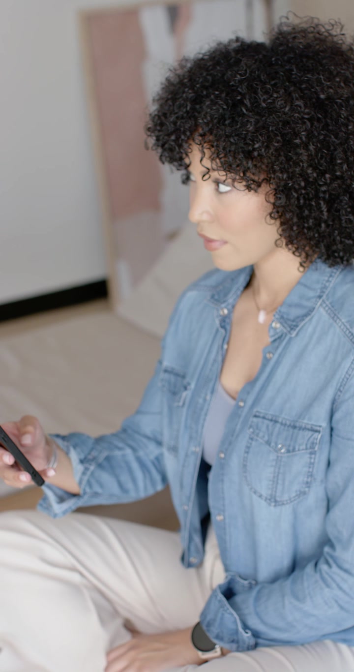 Video verticale: Donna afroamericana seduta su un tappeto a casa, che controlla il telefono e digita messaggi.