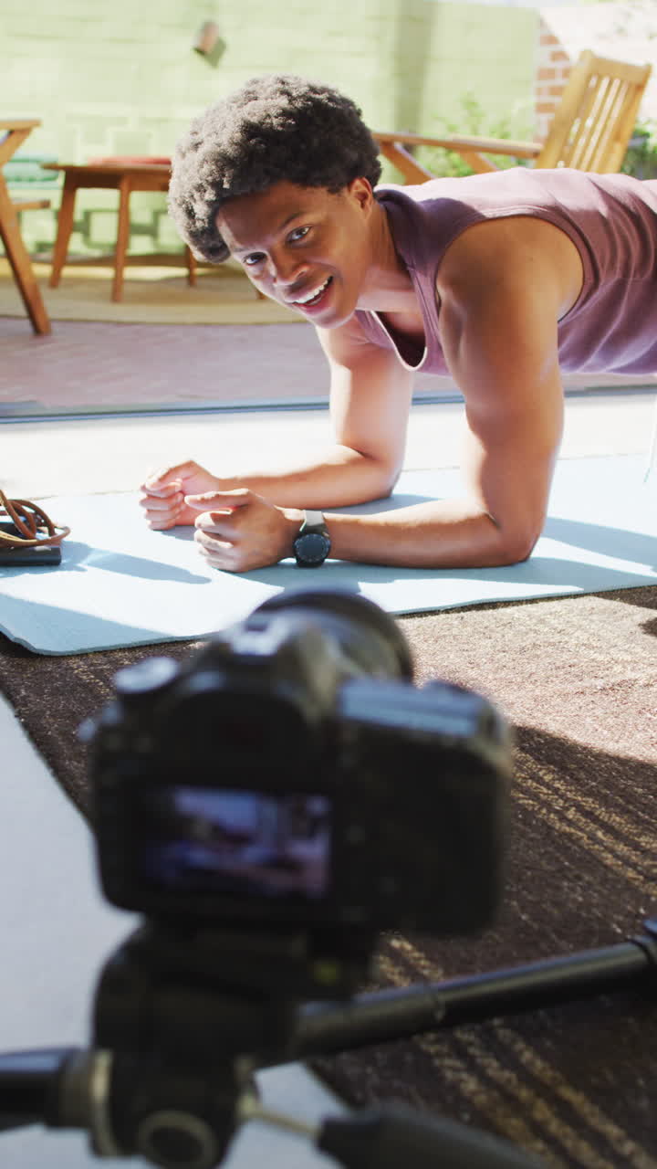 video vertical de vlogger masculino afroamericano haciendo ejercicio de tabla y filmando con cámara