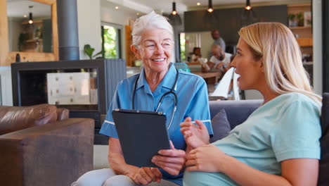 Ältere-Hebamme-Mit-Digitalem-Tablet-Besucht-Schwangere-Frau-Zu-Hause-Mit-Familie-Im-Hintergrund