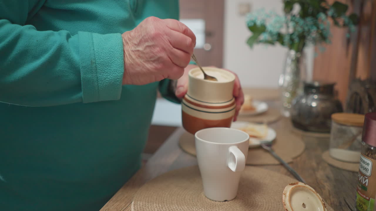 Elderly woman adding sugar to cup while preparing tea or coffee, cozy breakfast scene. Home kitchen with fresh flowers and simple table setting, highlighting homemaking and daily routine