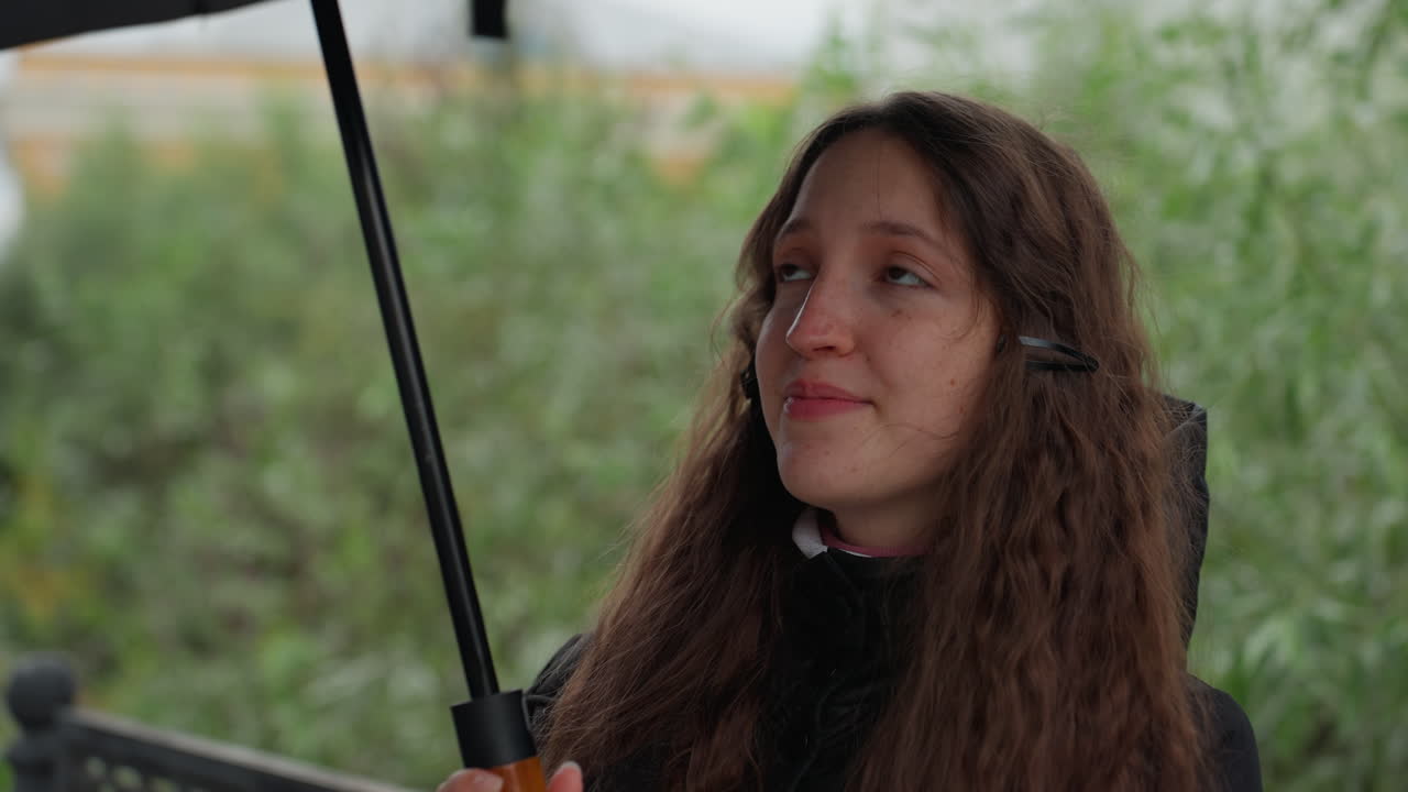 Mulher caucasiana caminhando com guarda-chuva, fone de ouvido e casaco com capuz passeando por um parque urbano nublado, close-ups capturam gotas no toldo preto, sorriso gentil e olhar contemplativo, outono