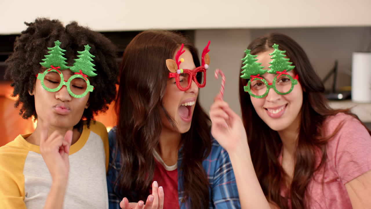 Women friends wearing festive glasses celebrating Christmas with candy canes by fireplace