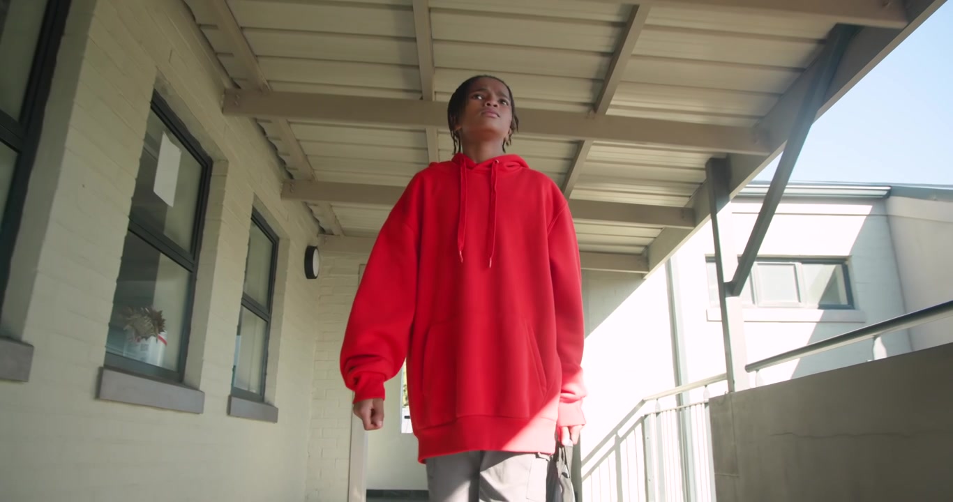 Remaja pria Afrika-Amerika berhoodie merah dari koridor, mendekati kamera sambil memegang tas dan melirik ke atas