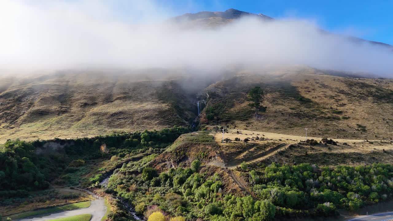Aerial footage captures a mist-covered mountain landscape with lush greenery and winding paths under clear blue skies