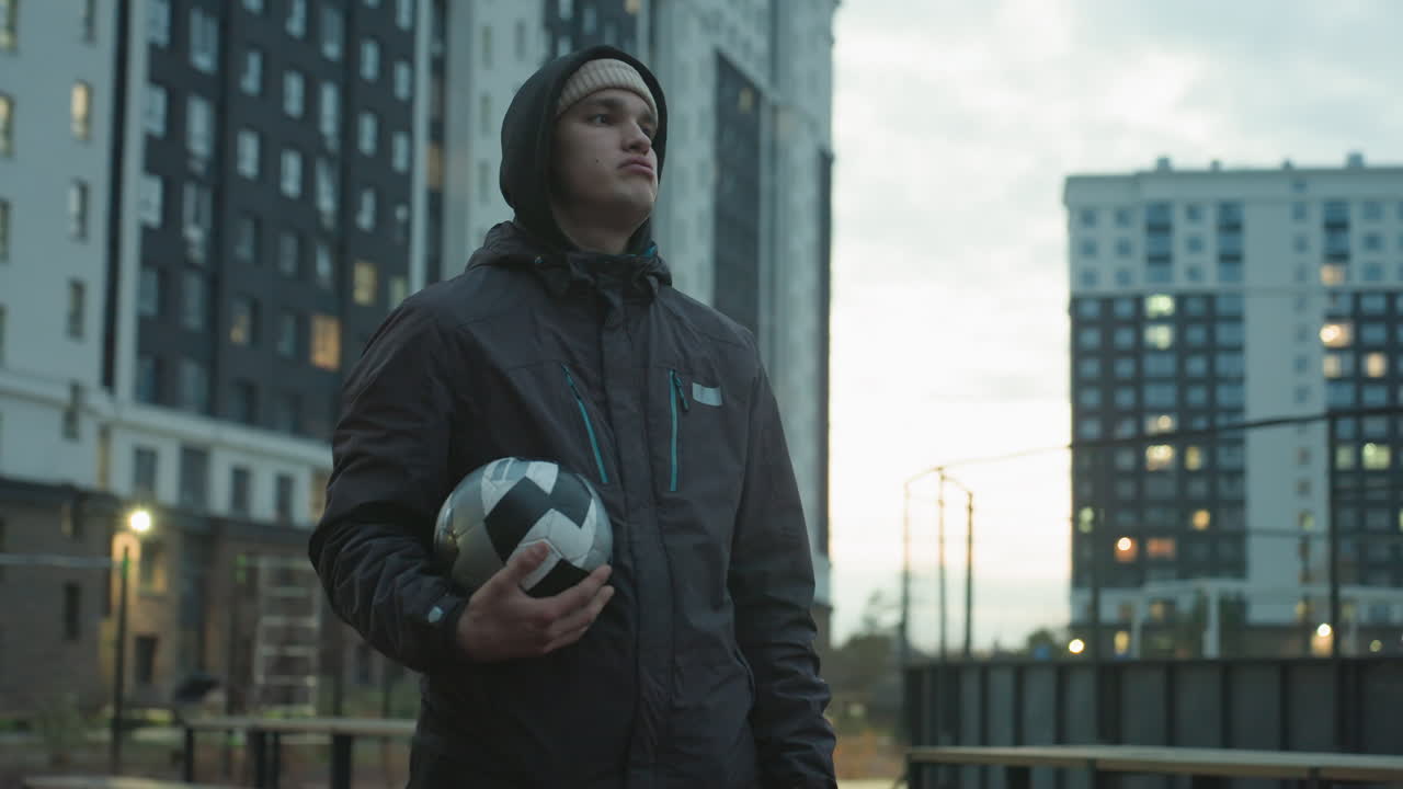 Man holding soccer ball standing confidently in modern sport arena surrounded by tall residential buildings under a cloudy evening sky
