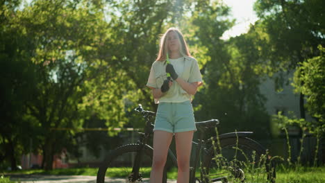 woman stands in sunlight, holding green air pump next to bicycle, using it to cool her face as her hair flutters gently in breeze, background highlights lush trees and distant building