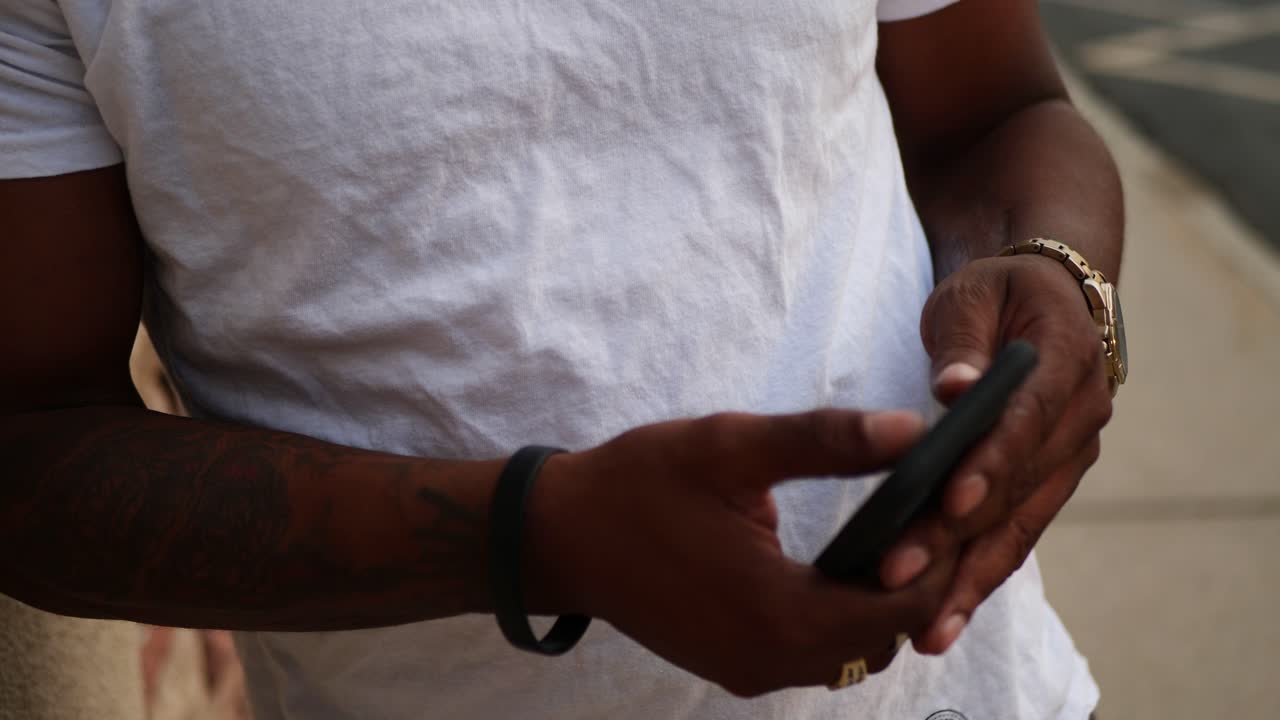 African American man on his phone texting and searching the internet in a white shirt and gold chain around his neck, next to brick and mortar building as camera pans up