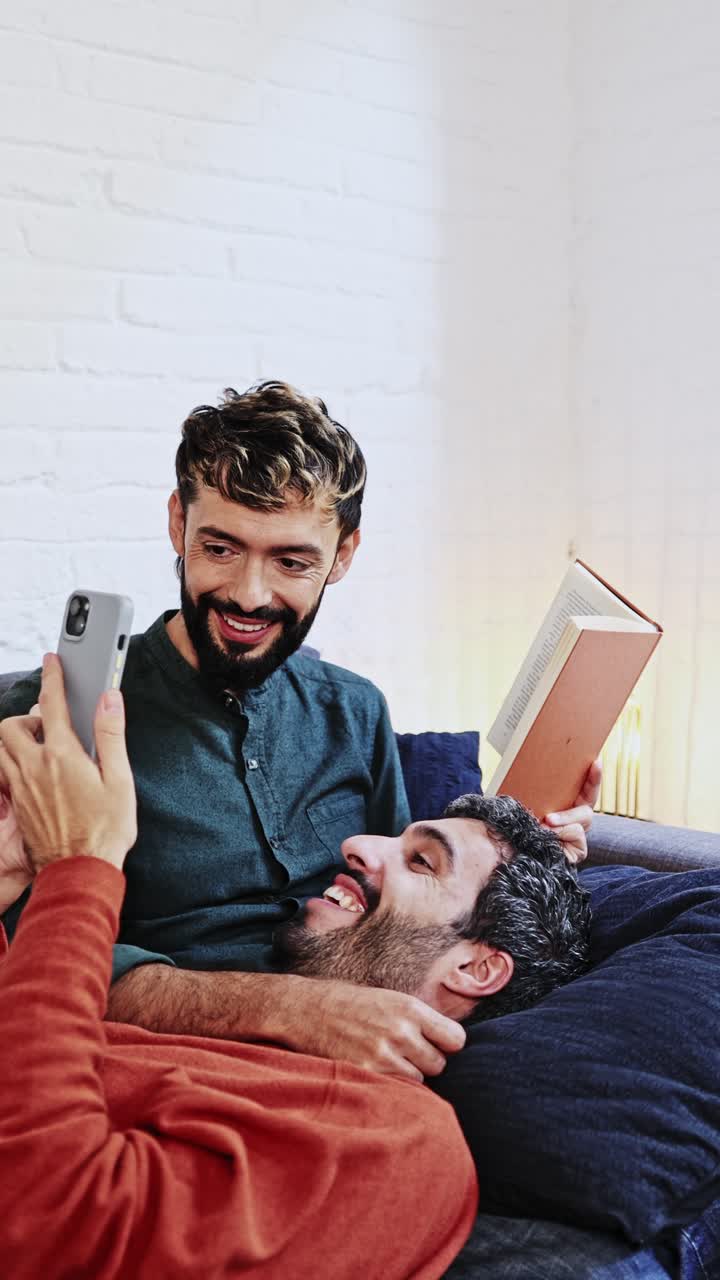 Coppia gay felice che si rilassa sul divano, guardando lo smartphone insieme