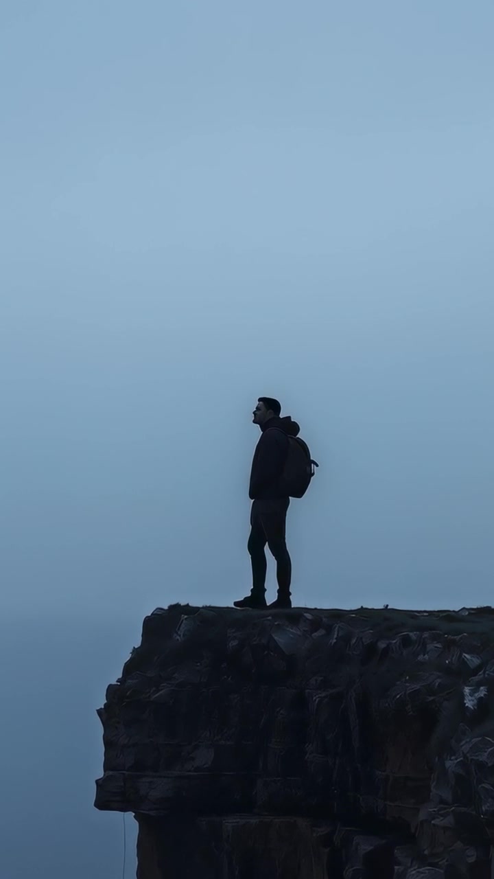 Video verticale: Un uomo con giacca con cappuccio raggiunge una scogliera, scrutando la nebbia in cerca di un percorso, con zaino e scarponi.