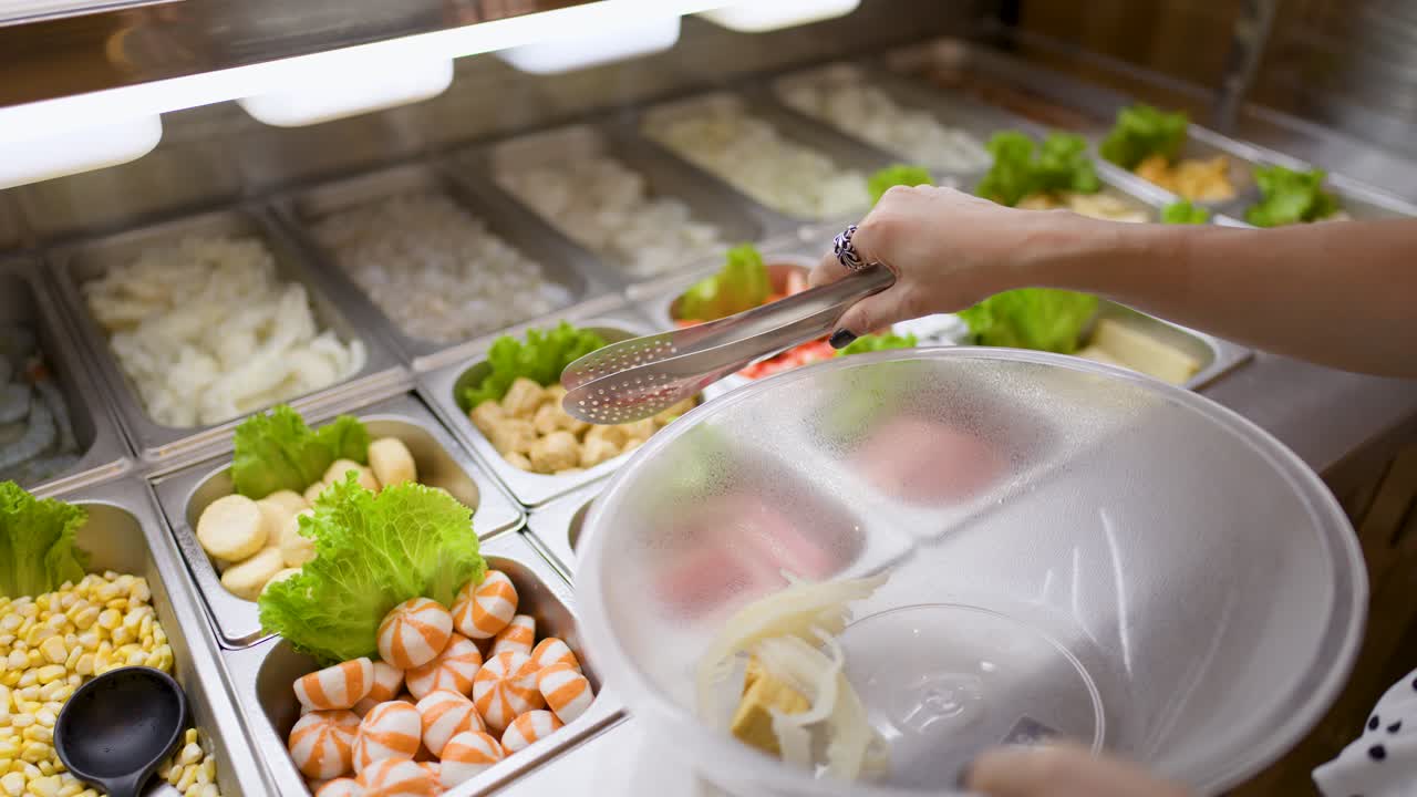 Person uses tongs to pick seafood and vegetables at well-lit buffet hotpot station