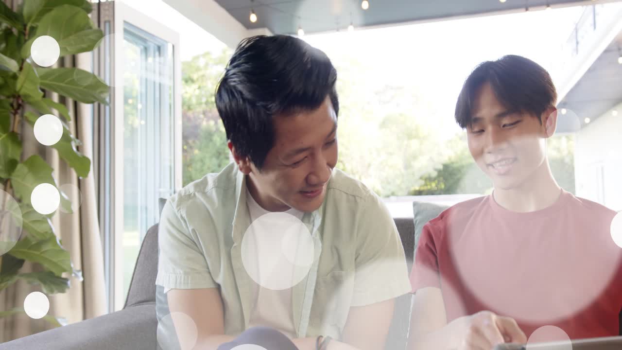 Father pointing at tablet, guiding son tapping tablet on couch, bokeh overlay enhancing learning