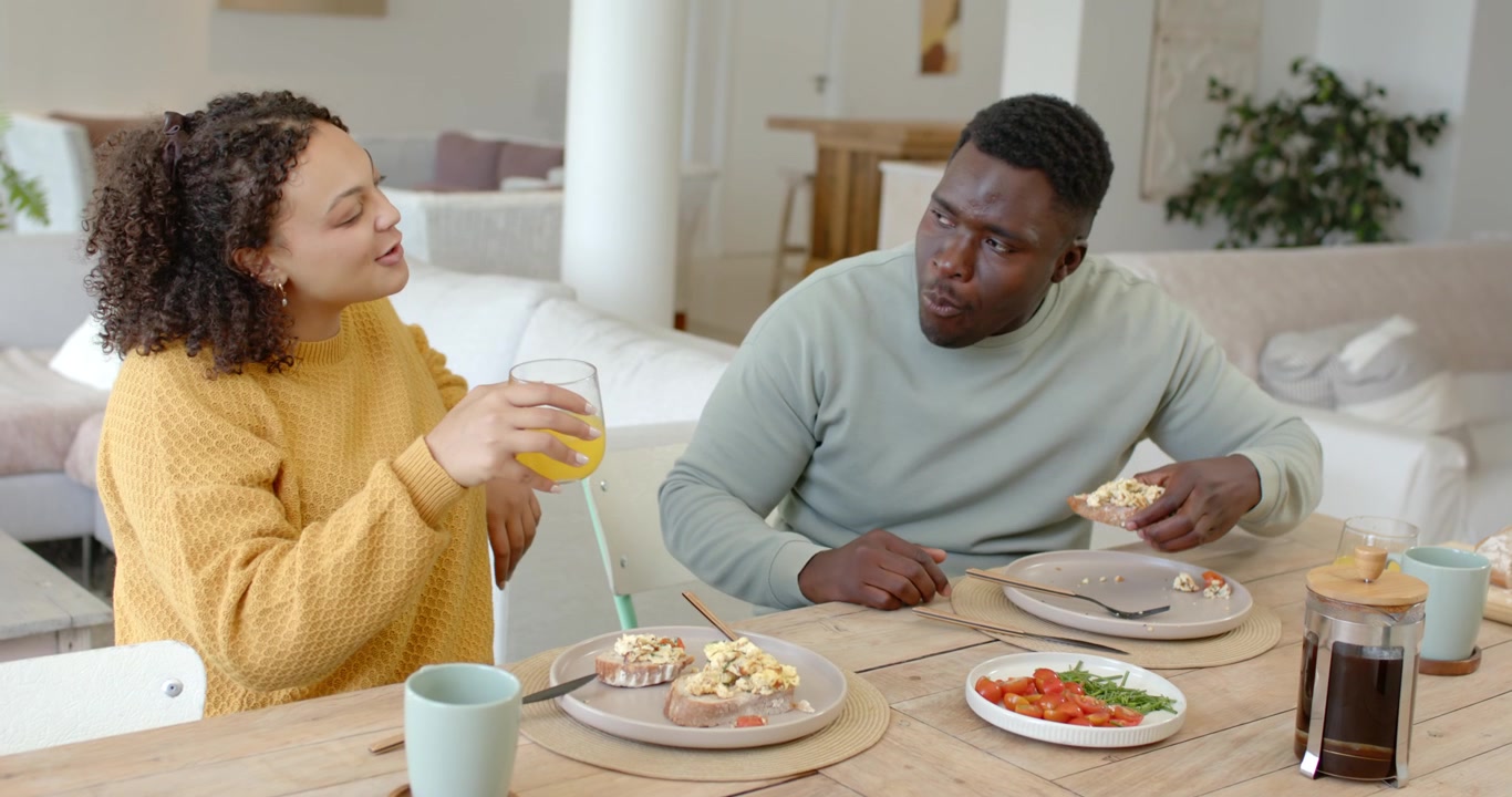 African American couple eating bagel at table, woman offering orange juice after man taking bite