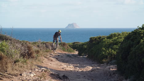 Caucasian-man-rides-mountain-bike-down-dirt-narrow-rocky-path-in-countryside-toward-blue-ocean-sea-with-Toro-islet-in-background,-Sardinia,-Italy,-static