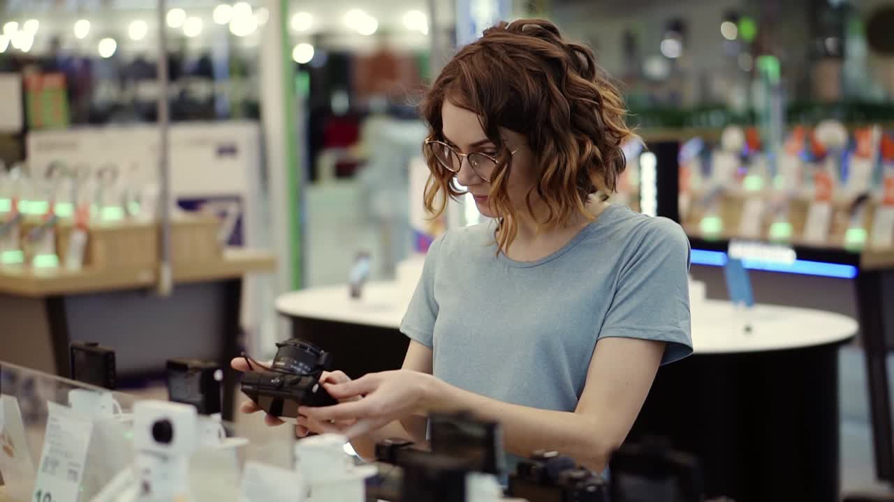 Young curly woman in glasses shopping for a new photo camera in the electronics store. Trying to decide on the best model. Have