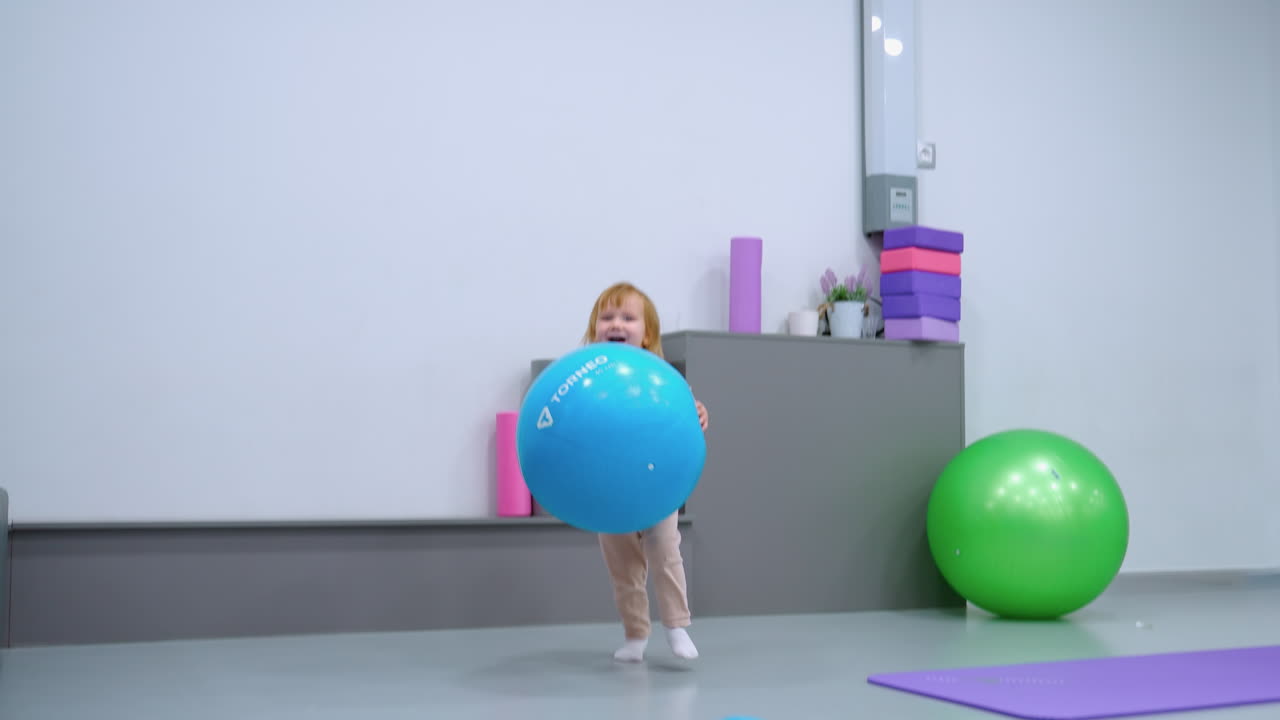 Balita bersemangat berlari dengan balon biru besar di studio kebugaran, dikelilingi oleh peralatan gym, termasuk bola latihan hijau, matras yoga, dan balok tumpuk