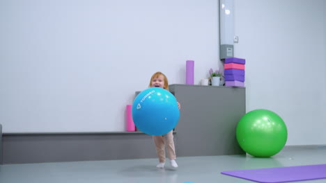 excited toddler running with large blue balloon in fitness studio, surrounded by gym equipment, including green exercise ball, yoga mats, and stacked blocks