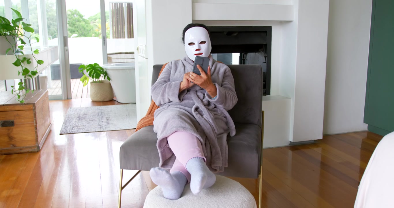 Senior woman sitting in gray chair tapping smartphone during facial mask routine at home