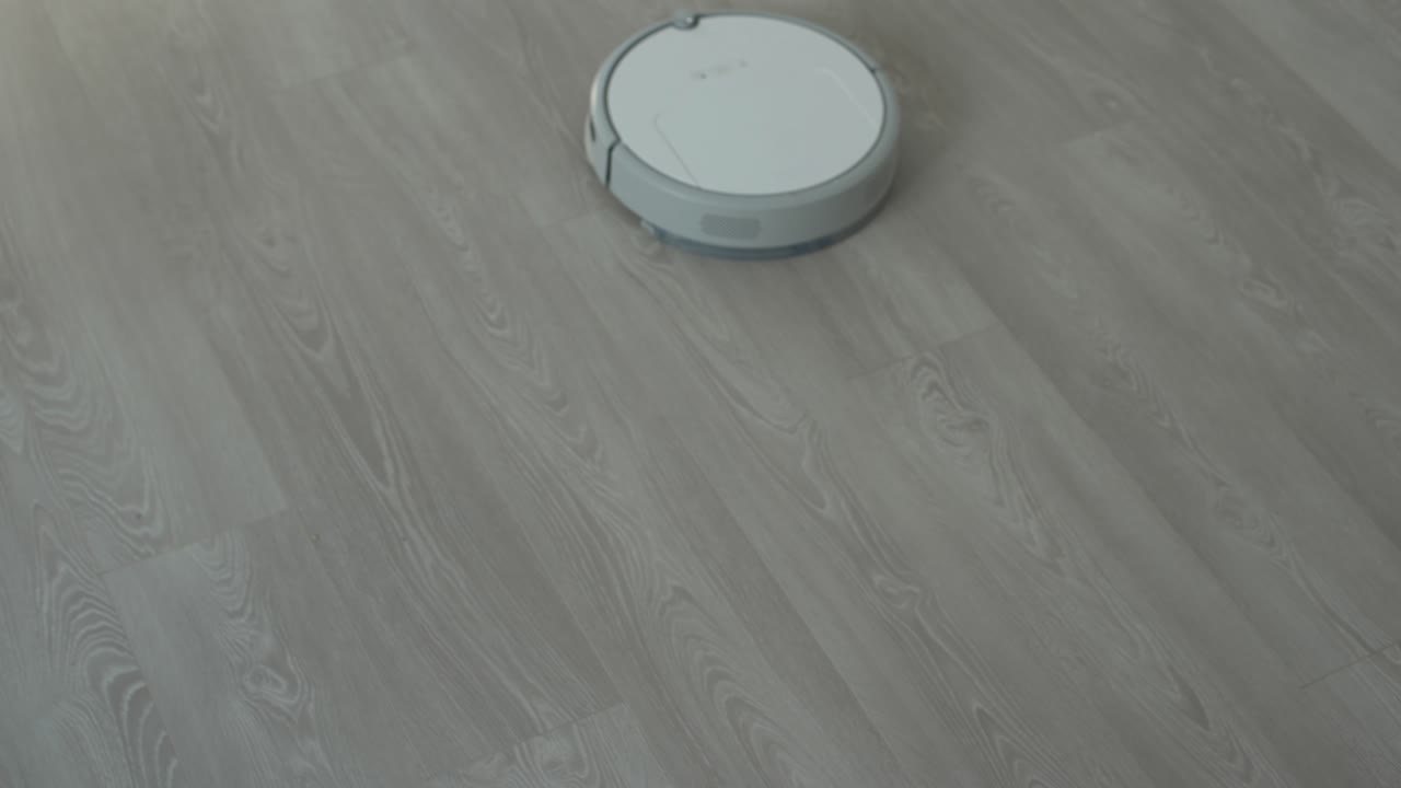 White robotic vacuum cleaner on laminate floor cleaning dust in living room interior. Smart housekeeping technology.