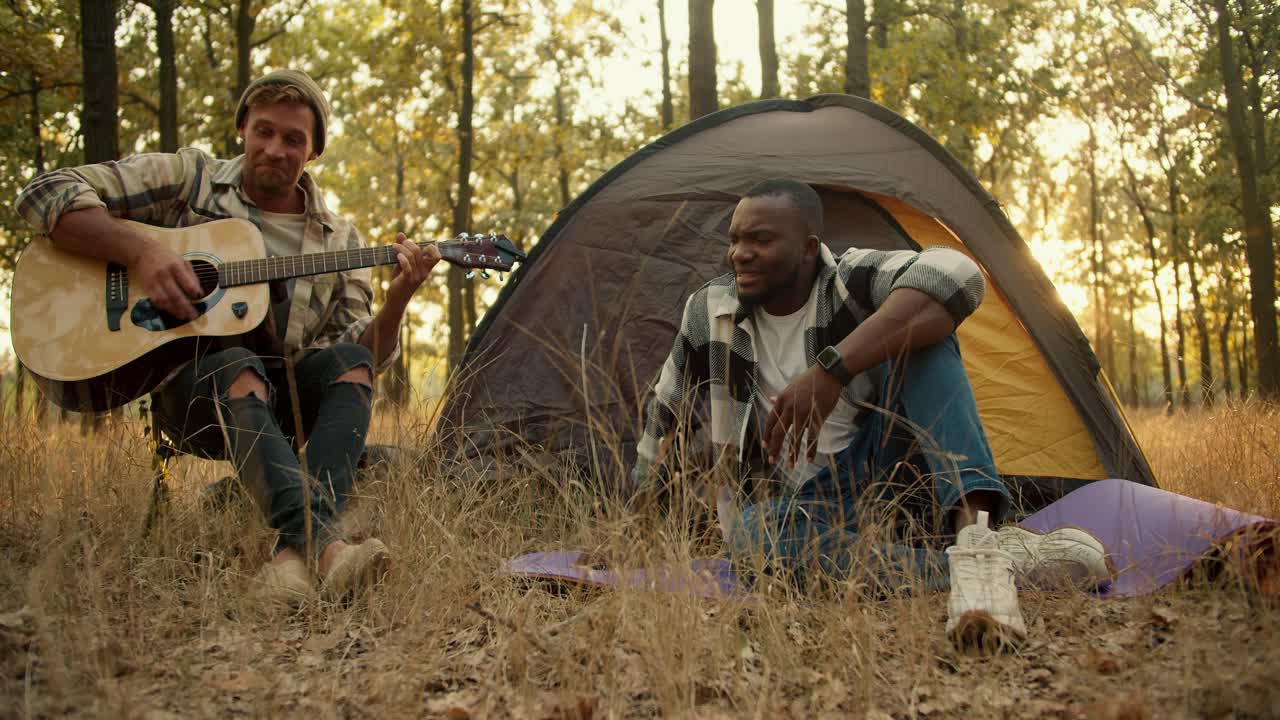 A happy man with black skin in a checkered shirt sings and a blond man in a hat plays the guitar against of a gray tent in a green forest during a summer halt. A happy company of two guys during a rest in a sunny light green summer forest