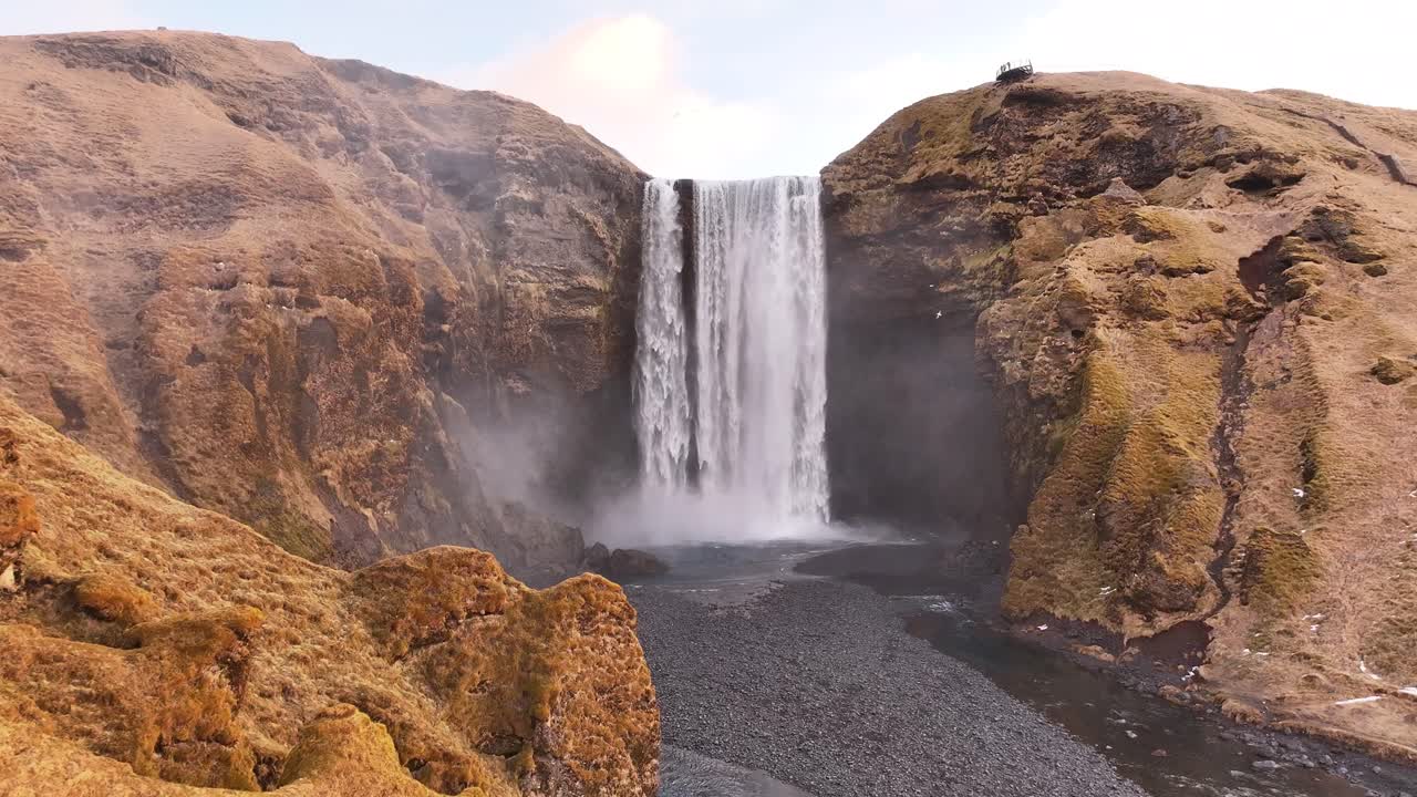 Aerial push-in toward Skógafoss waterfall in Iceland, mist rises from crashing water surrounded by mossy volcanic cliffs and rugged black pebbles, breathtaking view.