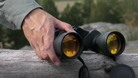 Man-picking-up-binoculars-on-fallen-tree-with-nature-hill-forest-wilderness-view