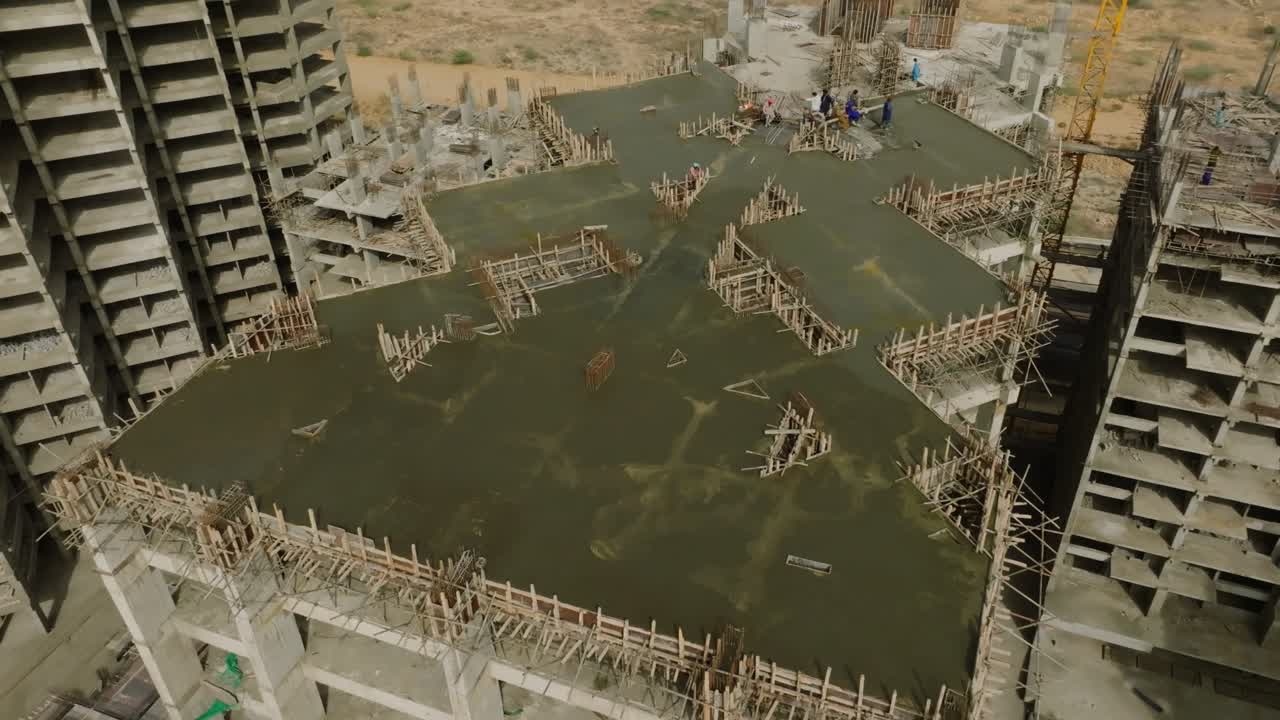 Drone captures an orbiting view of a partially completed high-rise in Karachi, where workers are seen on a freshly poured concrete slab surrounded by wooden scaffolding and exposed rebar structures.