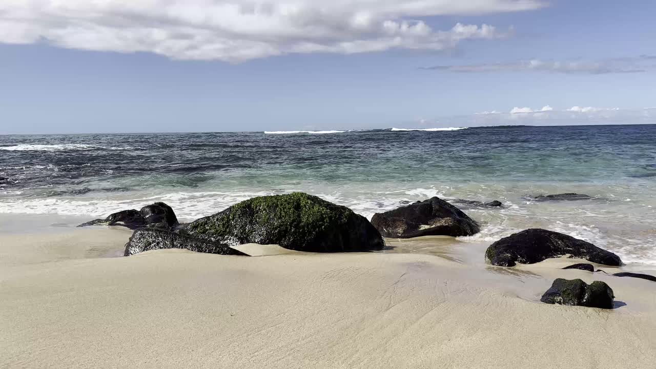 una tranquila playa hawaiana con ondas suaves golpeando contra rocas cubiertas de musgo bajo un cielo parcialmente nublado, mostrando la belleza natural de oahu
