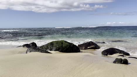 A-tranquil-Hawaiian-beach-with-gentle-waves-lapping-against-moss-covered-rocks-under-a-partly-cloudy-sky,-showcasing-Oahu's-natural-beauty