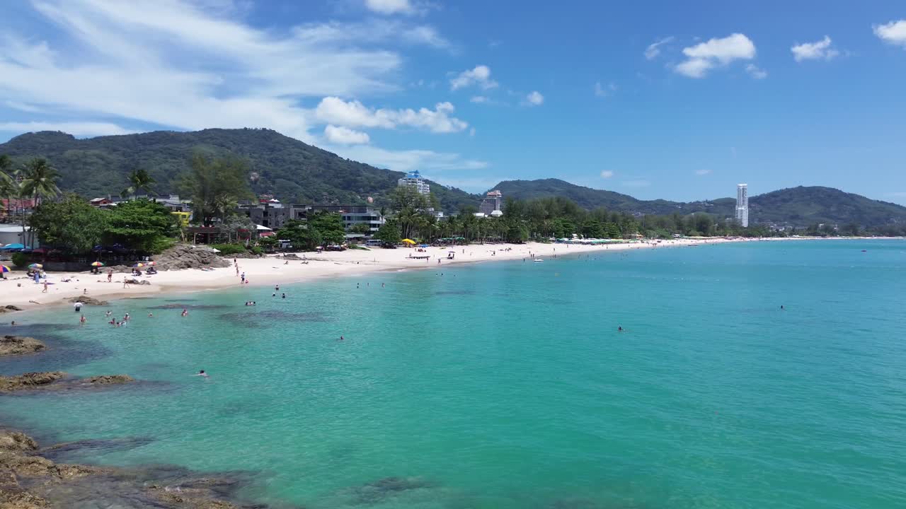 Smooth left-to-right drone shot over Patong Beach, showcasing colorful clear turquoise waters, white sand, and the lively Phuket coastline.