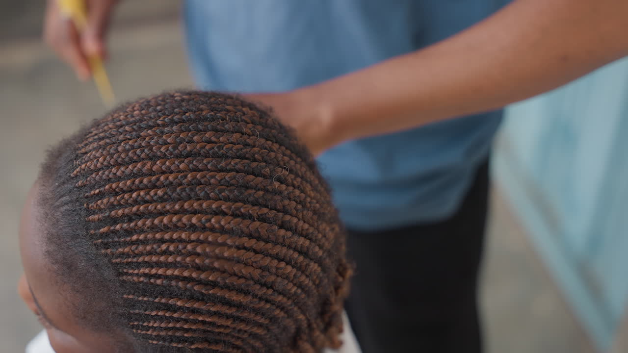 Primo piano di una ragazza nera che si fa fare le trecce cornrow. Le mani di una stilista nera intrecciano file strette sul cuoio capelluto, in un ambiente tranquillo sul portico, con luce naturale. Dettagliata texture dei capelli, movimenti pazienti e attenti, cura familiare intima.