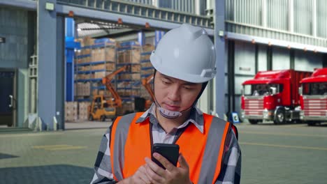 close up of asian male engineer with safety helmet using smartphone and looking around while standing , outside of logistics distributions warehouse