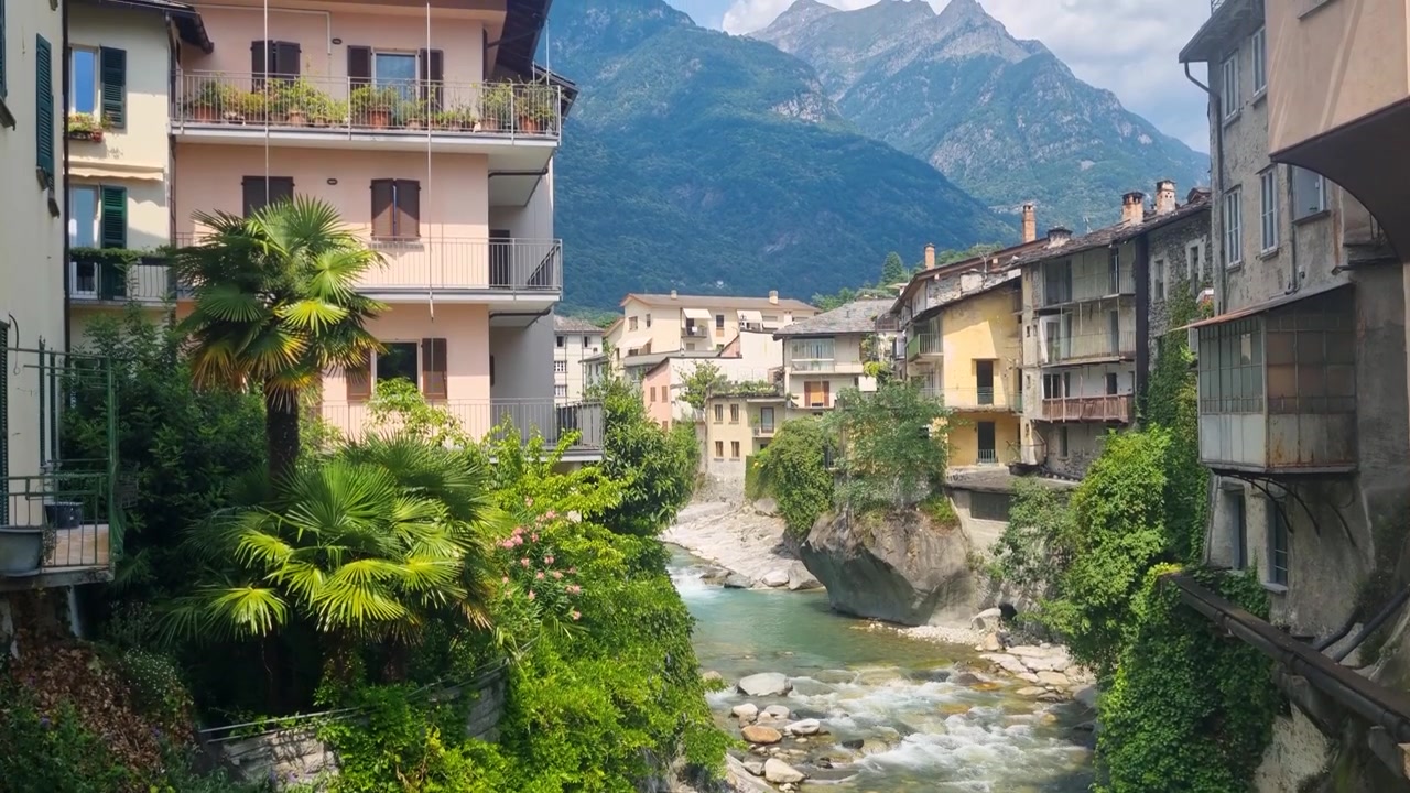Vista pittoresca di Chiavenna, Lombardia, Italia, con edifici colorati lungo il fiume Mera, vegetazione mediterranea e le aspre Alpi Retiche, che mette in mostra questo storico villaggio alpino.