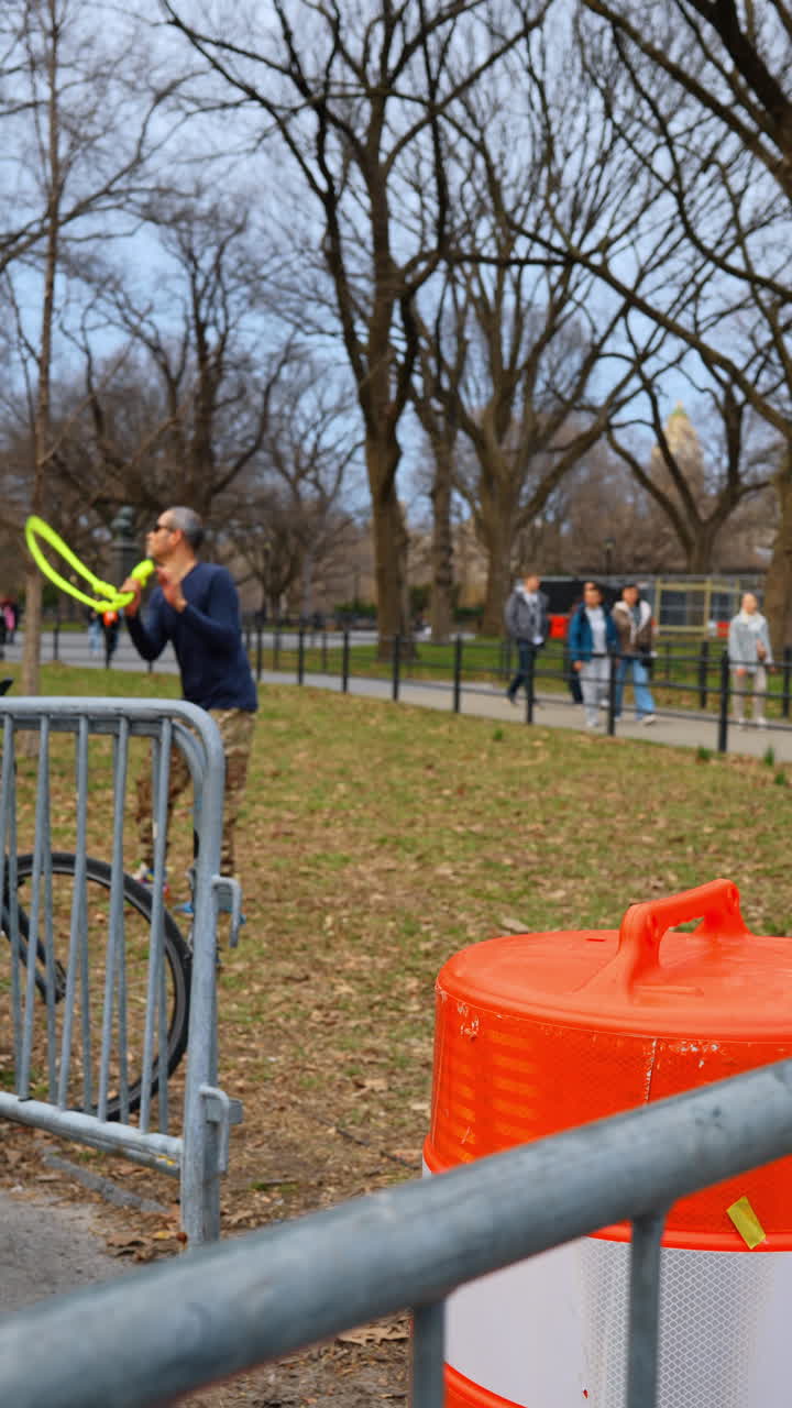 Nowy Jork, USA, 15 kwietnia 2026: Pomarańczowa beczka drogowa obok barykady w Central Parku i artysty ulicznego. Pomarańczowa beczka drogowa i metalowa barykada na pierwszym planie, artysta uliczny i przechodnie rozmyci w tle.