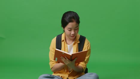 asian woman student with a backpack holding and reading a book while sitting in the green screen background studio