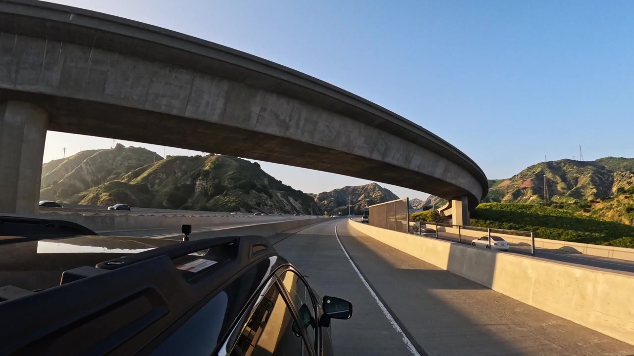 ロサンゼルスの405号線北行きで撮影された、魅惑的な走行中の車の映像。誰もいない空の高速道路、晴れた山の風景、高速道路のジャンクションの陸橋、そしてレンズに差し込む太陽のフレア