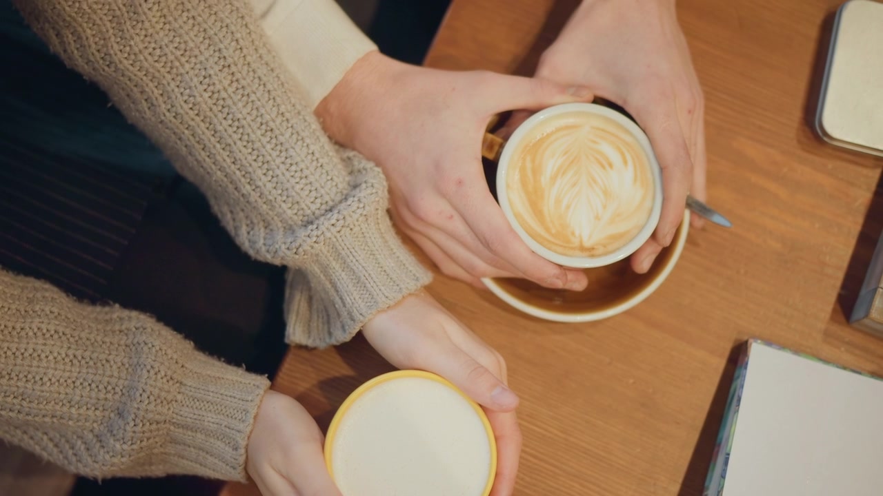 Due donne tengono tazze di caffè su un tavolo di legno, una con latte art e l'altra piena di latte. Entrambe indossano maglioni di lana beige e dei quaderni sono visibili nelle vicinanze. L'illuminazione soffusa crea un ambiente interno caldo.