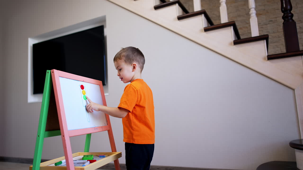 Serious toddler boy in orange t-shirt stands looking at the whiteboard. Kid comes up to board and touches the colorful magnets.
