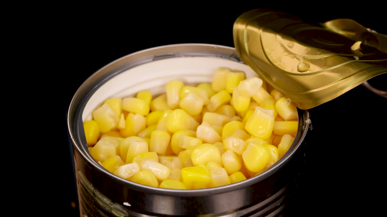 Cooked corn kernels fall into an open metal can against a black background, captured in close-up with bright, even lighting and minimal camera movement