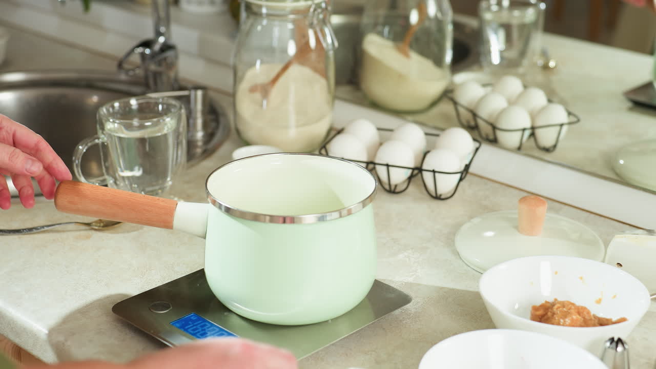 Partial view of lady placing modern pastel pot with wooden handle on digital kitchen scale, pressing button to activate weighing, surrounded by kitchen items including eggs and bowls, jar of flour