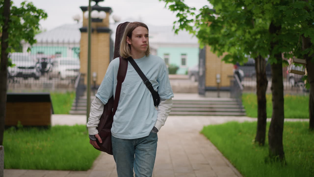 Mladý kytarista kráčí parkem s pouzdrem na kytaru přes rameno, má na sobě ležérní džínovinu, dlouhé blond vlasy, zamyšlený výraz, lavičky a listnaté stromy, náměstí v pozadí, uvolněná indie atmosféra, ranní světlo