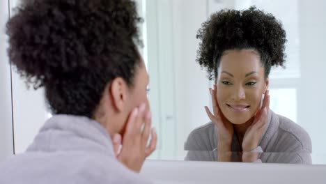 Happy-biracial-woman-in-bathrobe-looking-in-mirror-in-bathroom,-slow-motion