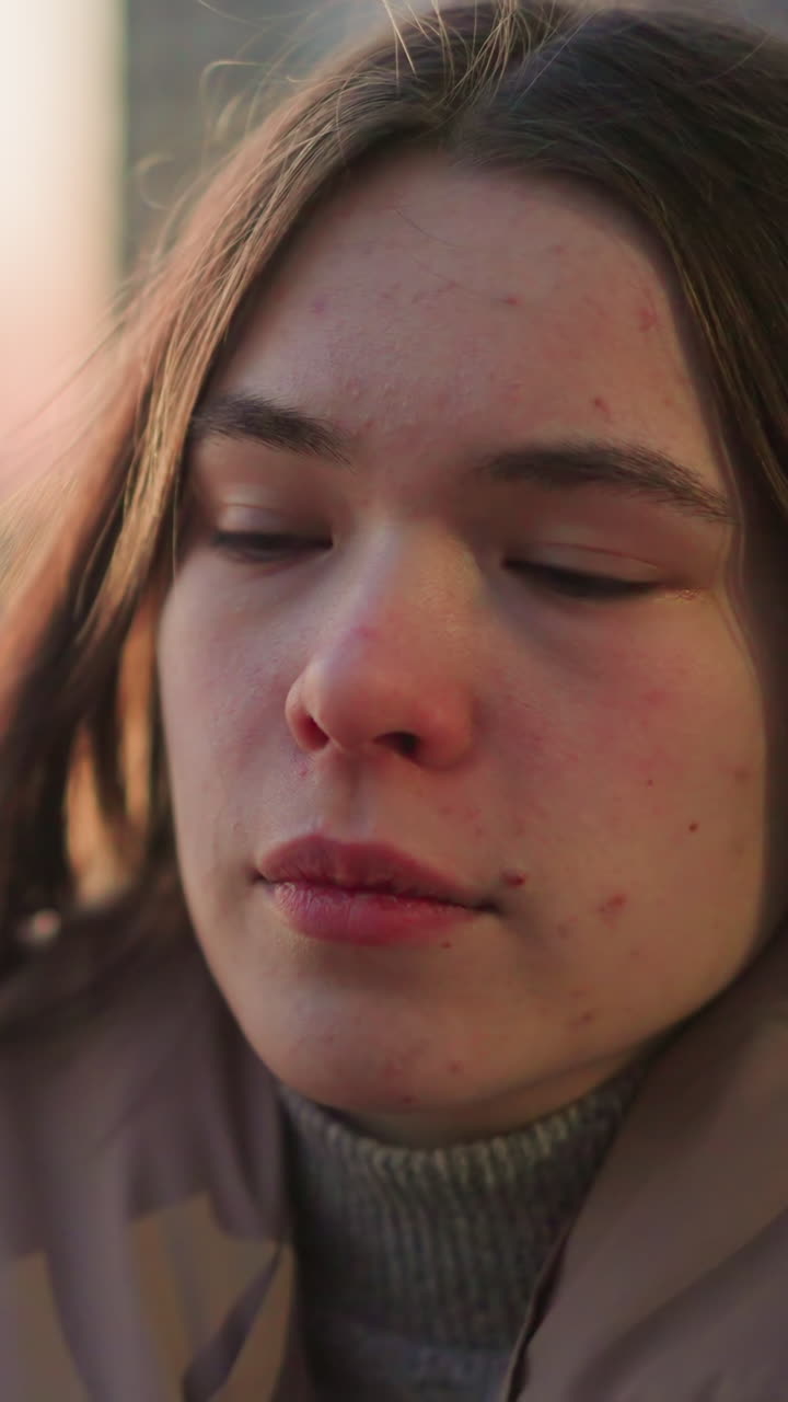un primer plano de una mujer joven con una chaqueta de melocotón, que parece perdida en profundos pensamientos con una débil sonrisa en su rostro. el fondo urbano borroso y la suave luz de la tarde