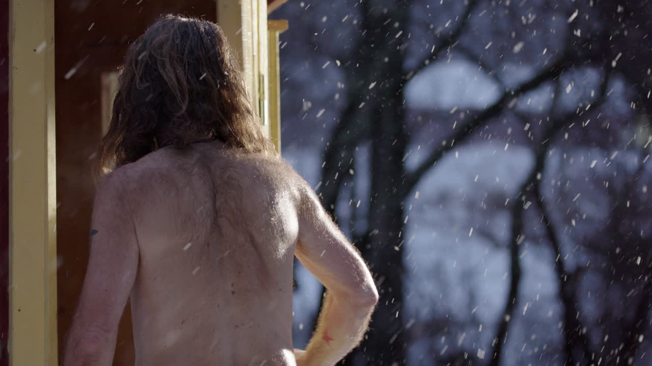 Spiritual bare chested scraggy man standing in snowfall enters steaming sauna