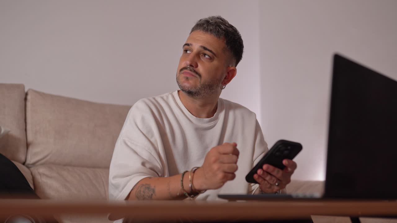 Man seated on sofa while typing on smartphone with serious expression, working next to open laptop on coffee table in modern living room